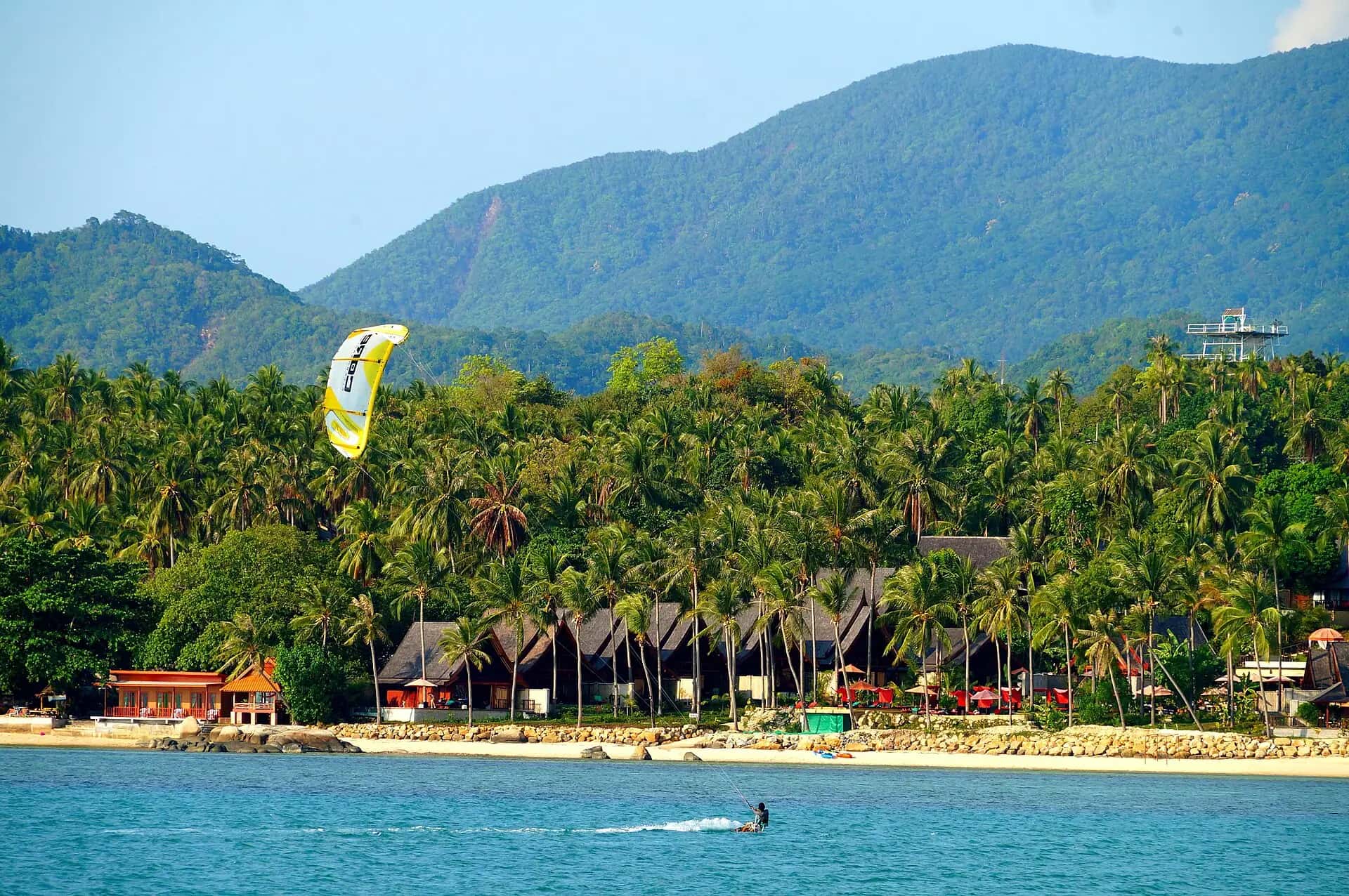 A kitesurfer gliding past a palm-lined beach with jungle mountains behind